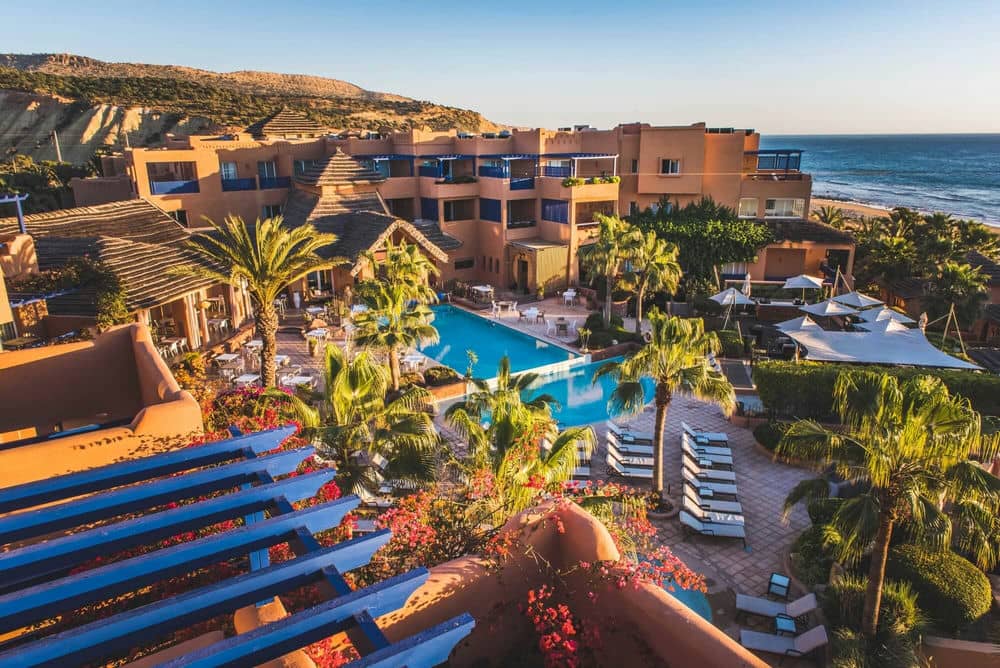 http://Vue%20aérienne%20d’un%20hôtel%20balnéaire%20au%20style%20marocain%20avec%20piscine,%20palmiers%20et%20plage%20à%20l’horizon.