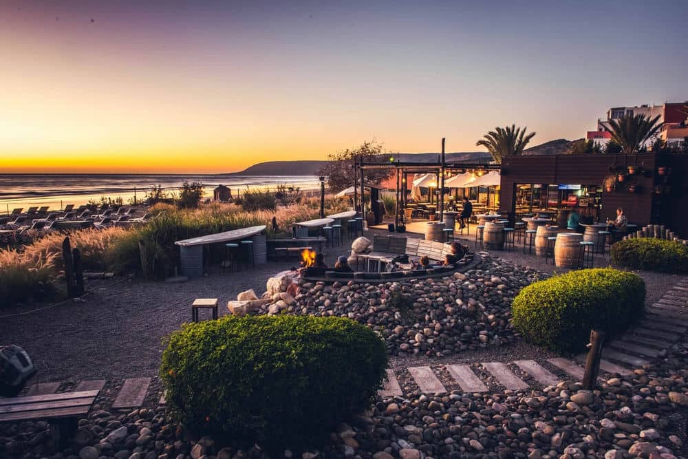 Restaurant de plage avec feu de camp, végétation et vue sur l’océan au coucher du soleil.