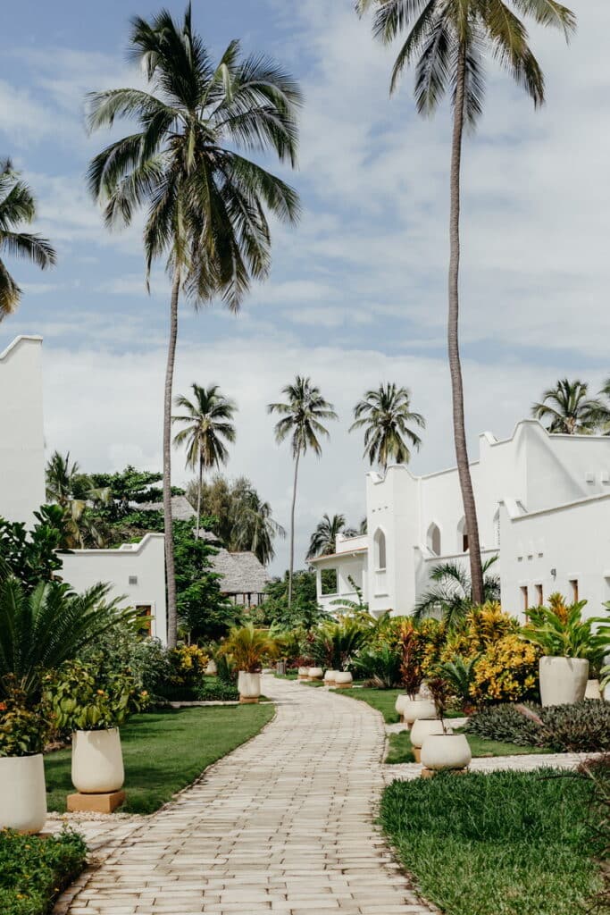 Allée pavée bordée de plantes exotiques et de bâtiments blancs de style swahili dans une station balnéaire tropicale.