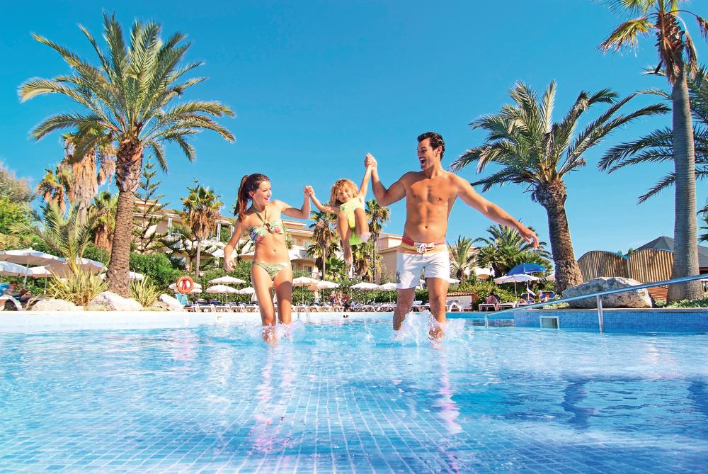 http://Famille%20avec%20enfant%20jouant%20dans%20une%20piscine%20peu%20profonde,%20entourée%20de%20palmiers%20et%20de%20transats.
