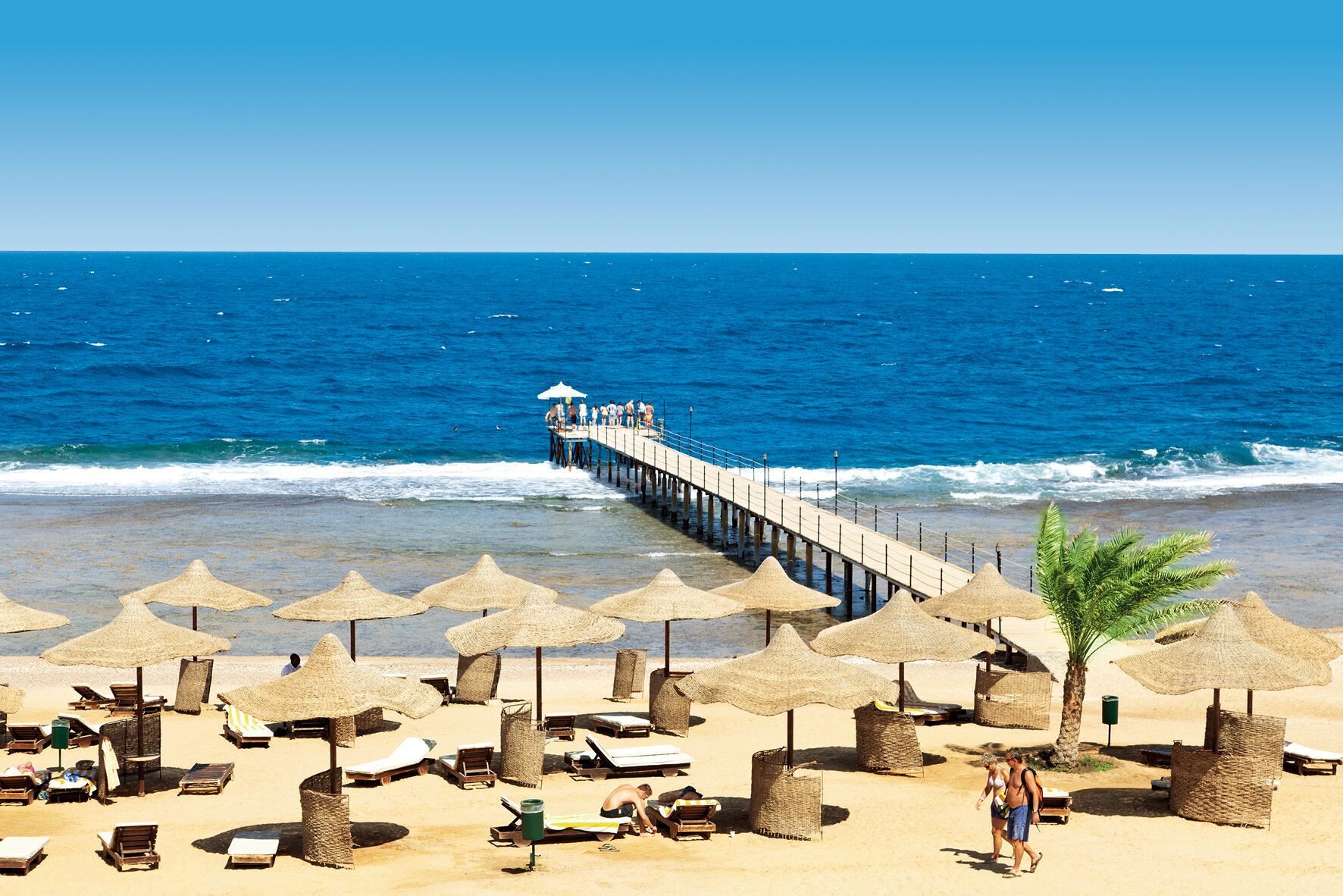 http://Plage%20aménagée%20avec%20parasols%20en%20paille,%20chaises%20longues%20et%20ponton%20s’avançant%20sur%20la%20mer.