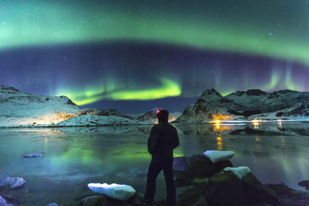 Personne observant les aurores boréales illuminant le ciel nocturne au-dessus de montagnes enneigées.