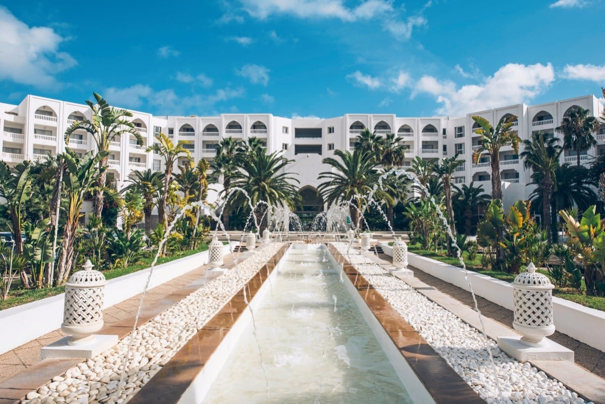 http://Hôtel%20de%20luxe%20à%20l’architecture%20blanche%20entouré%20de%20palmiers%20et%20d’une%20allée%20avec%20fontaines%20en%20Tunisie.
