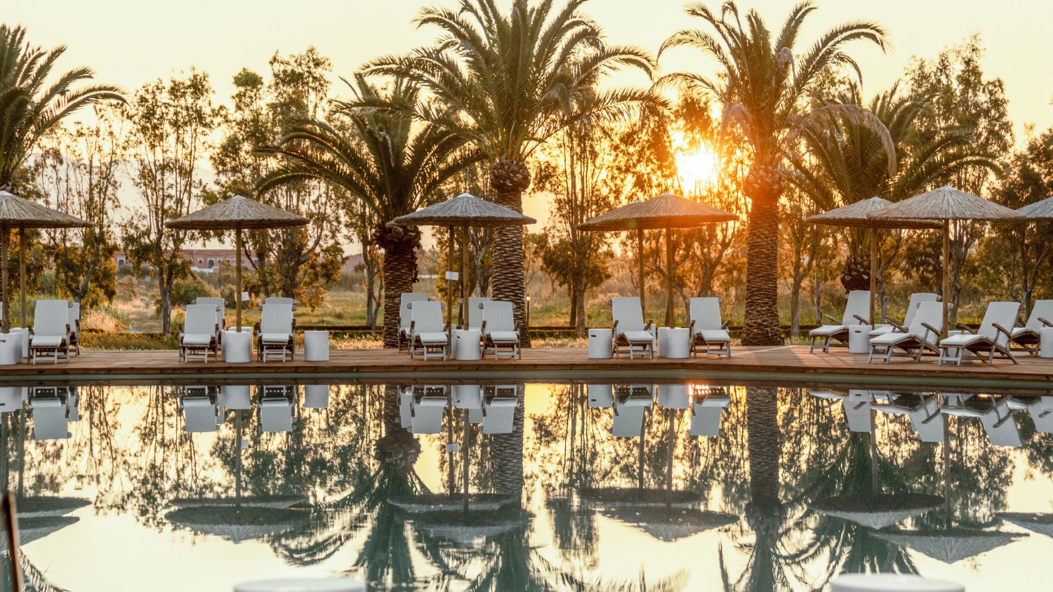 http://Piscine%20extérieure%20avec%20transats%20et%20parasols,%20bordée%20de%20palmiers%20au%20coucher%20du%20soleil.