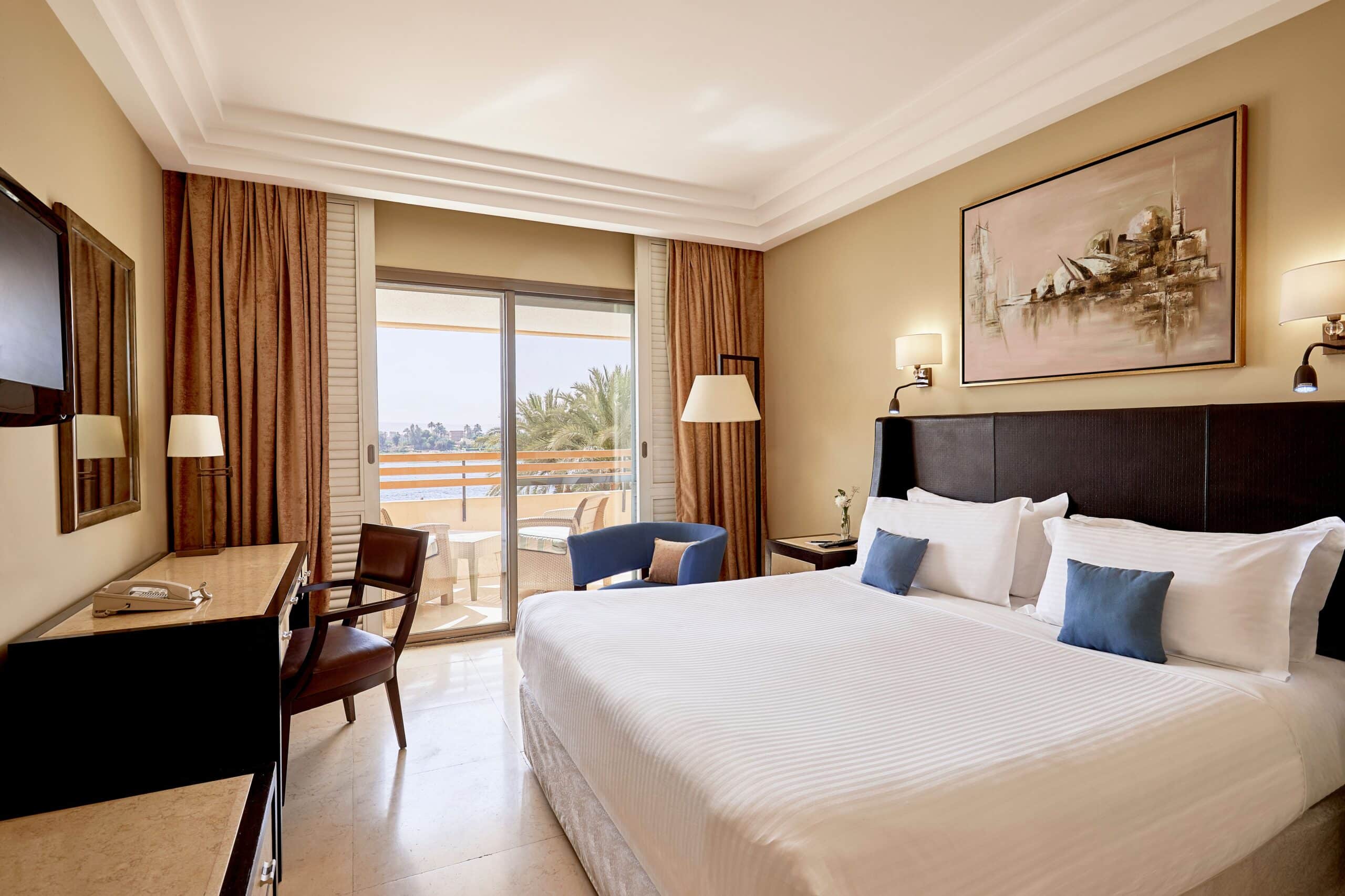 http://Chambre%20d'hôtel%20lumineuse%20avec%20un%20lit%20double,%20une%20décoration%20élégante,%20un%20balcon%20offrant%20une%20vue%20sur%20des%20palmiers%20et%20le%20fleuve.