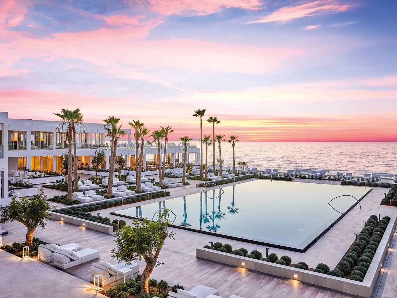 http://Piscine%20miroir%20au%20Grecotel%20avec%20vue%20directe%20sur%20la%20mer,%20bordée%20de%20palmiers,%20sous%20un%20ciel%20rose%20de%20coucher%20de%20soleil.