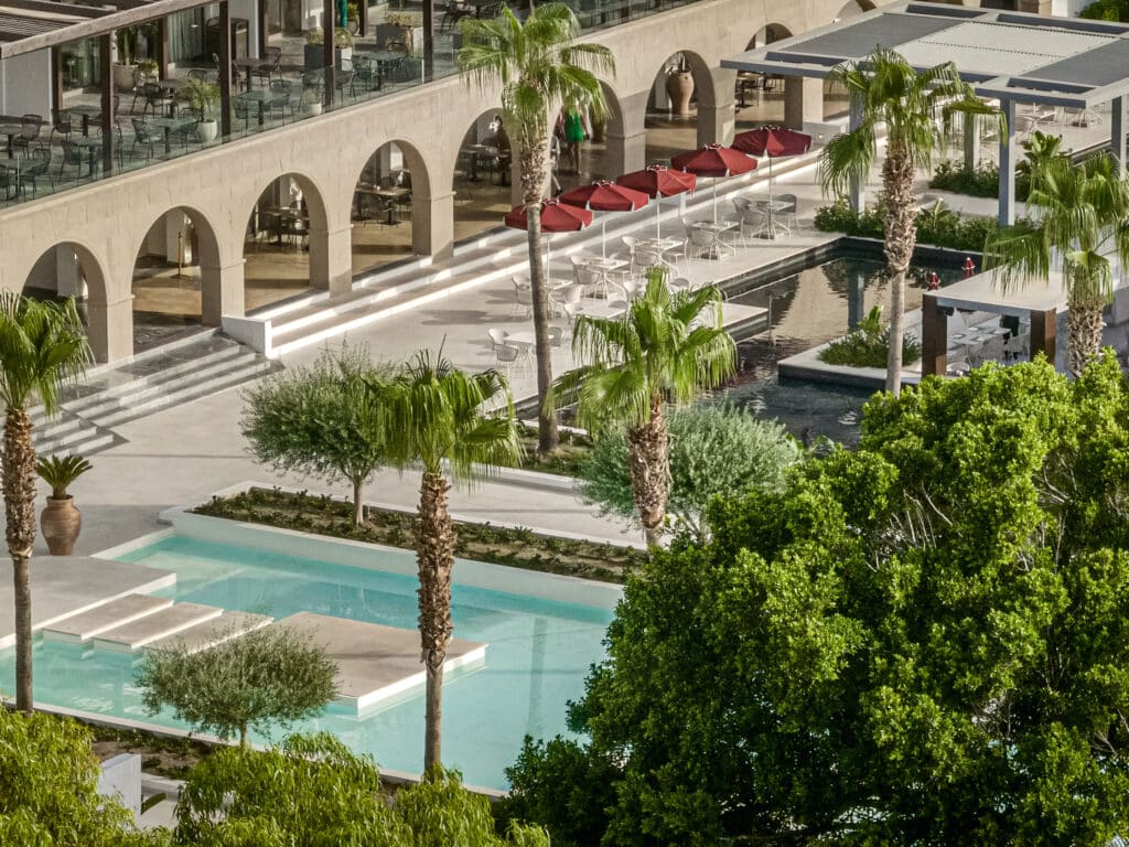 Terrasse d’hôtel avec arcades en pierre, bassins décoratifs, parasols rouges et palmiers.