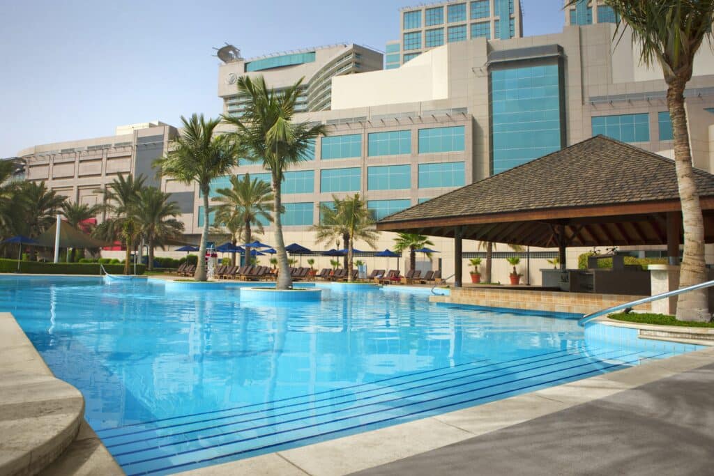 Grande piscine extérieure entourée de palmiers et d’un bar avec un hôtel moderne en arrière-plan.