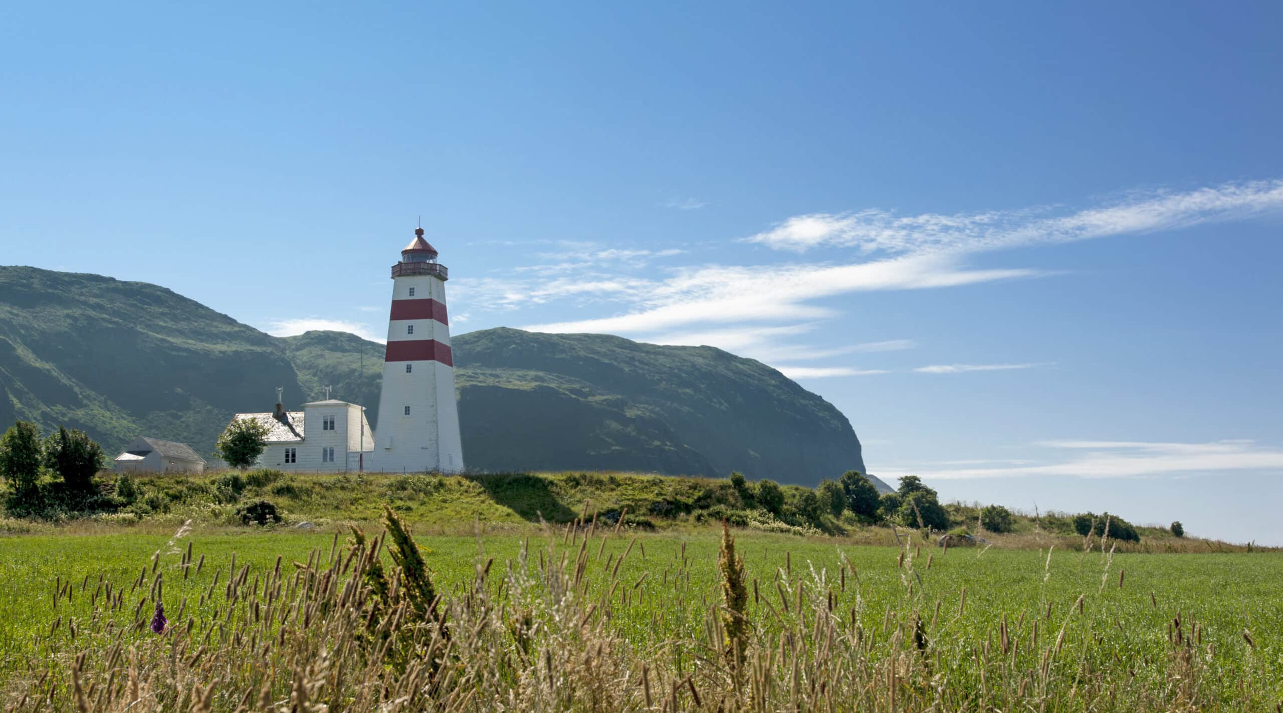 http://Phare%20blanc%20et%20rouge%20typique%20de%20la%20Norvège,%20perché%20sur%20une%20colline%20verdoyante%20avec%20des%20montagnes%20en%20arrière-plan%20sous%20un%20ciel%20bleu.