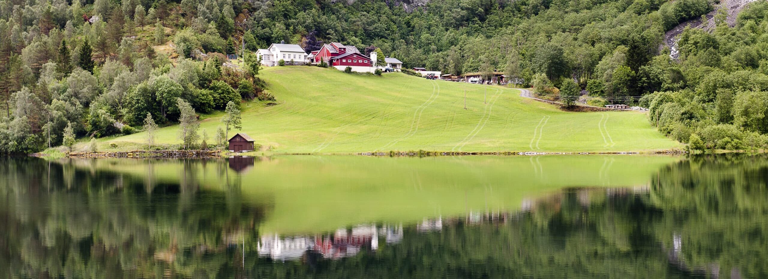 http://Reflet%20d’une%20ferme%20traditionnelle%20norvégienne%20sur%20les%20eaux%20calmes%20d’un%20fjord,%20entourée%20de%20montagnes%20verdoyantes.