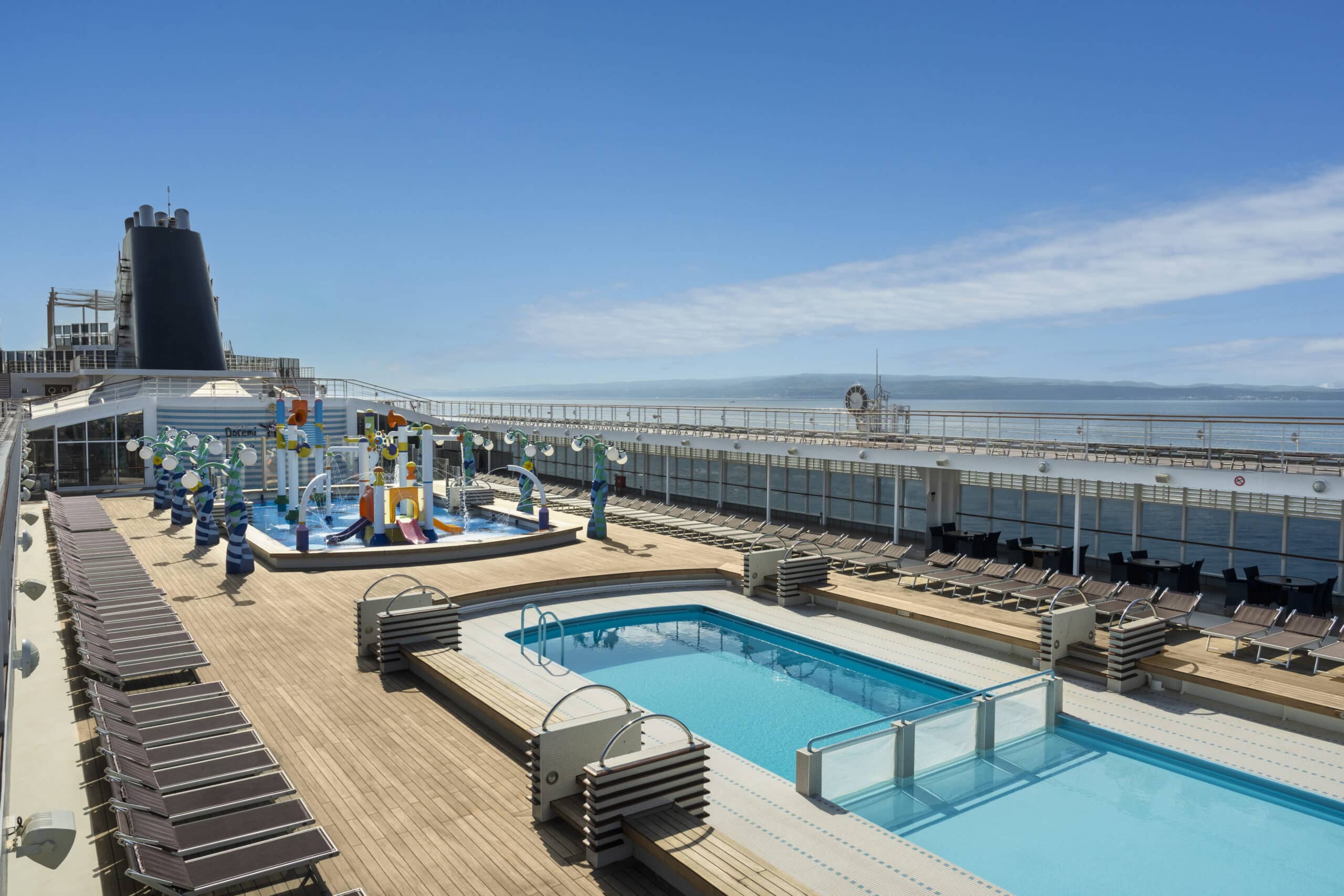 Deux piscines extérieures et une aire de jeux aquatiques pour enfants sur le pont d'un navire de croisière, avec transats et vue sur la mer.