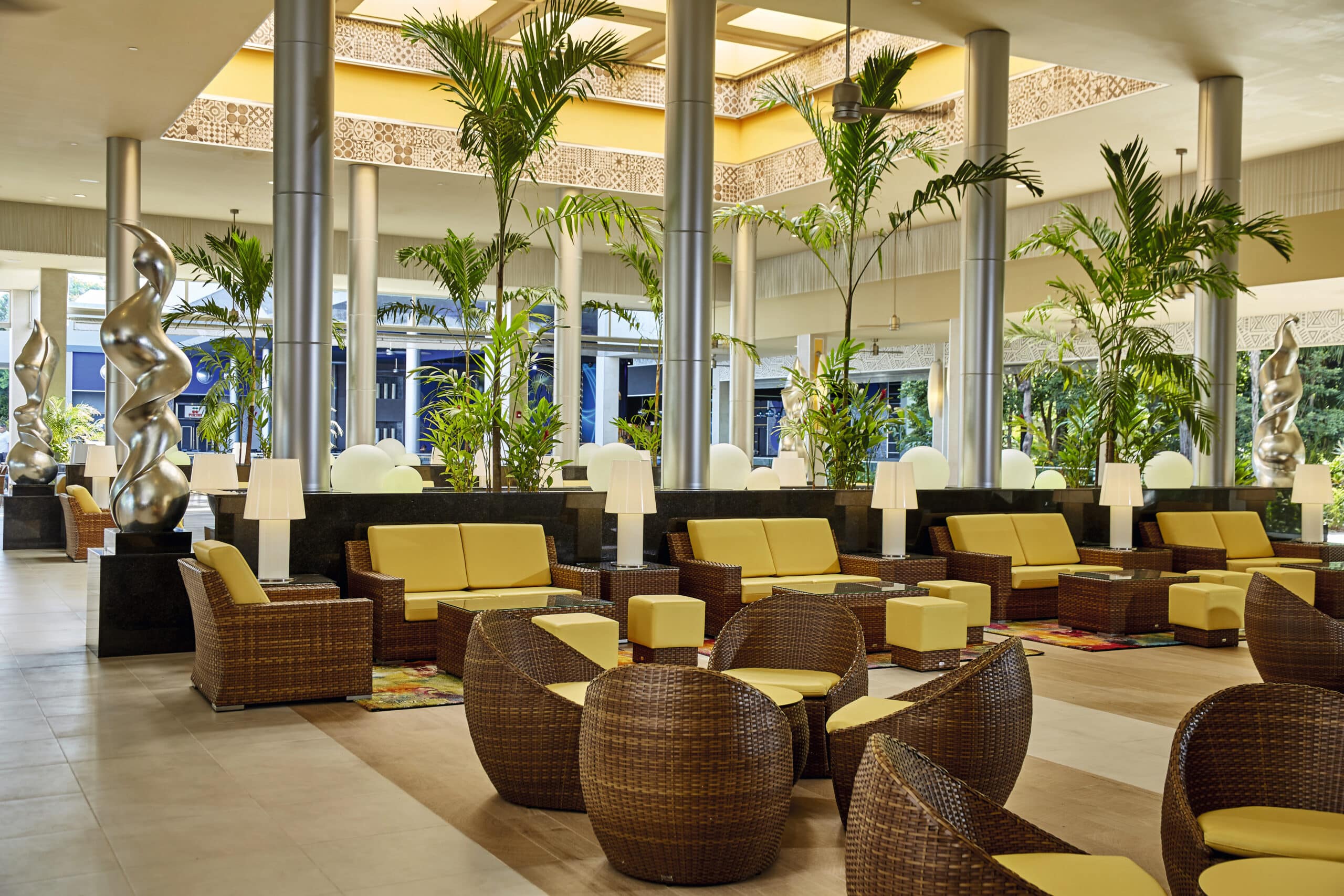 http://Hall%20d’hôtel%20lumineux%20avec%20mobilier%20en%20rotin,%20coussins%20jaunes%20et%20plantes%20tropicales.