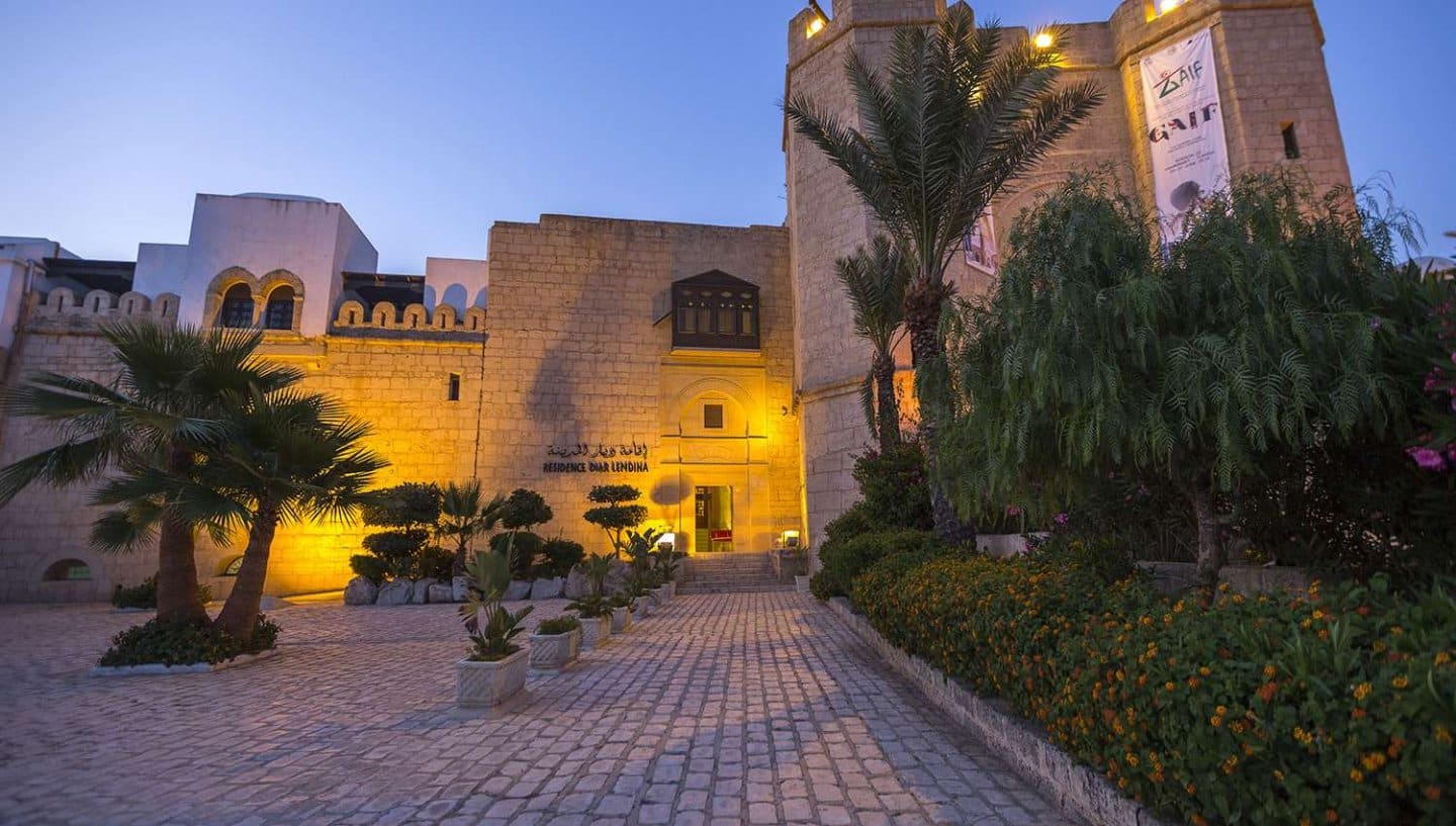 http://Entrée%20d’un%20hôtel%20de%20style%20tunisien%20éclairée%20à%20la%20tombée%20de%20la%20nuit%20avec%20palmiers%20et%20végétation