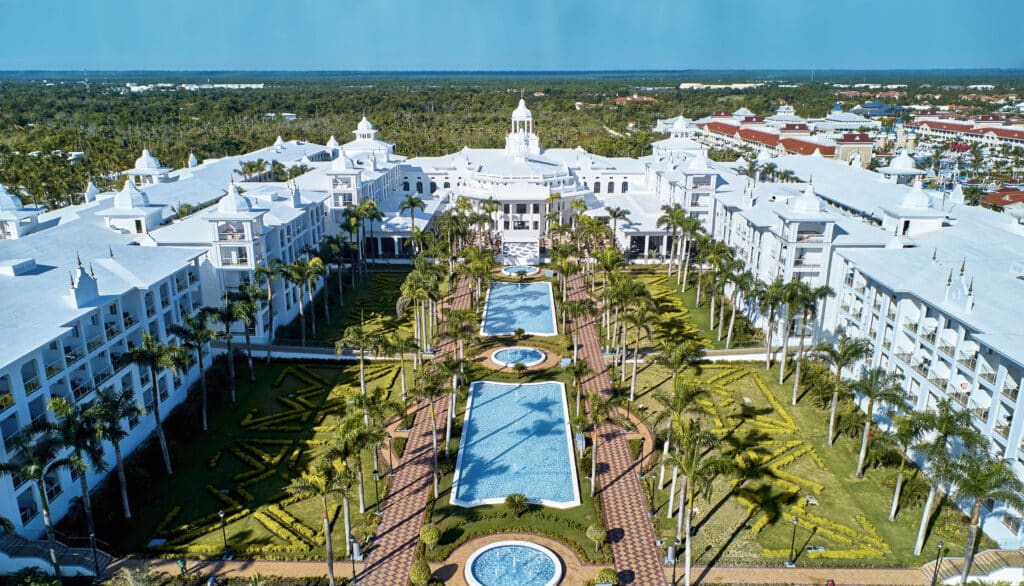 Vue aérienne d’un hôtel de luxe avec bâtiments blancs, jardins paysagers et piscines alignées.