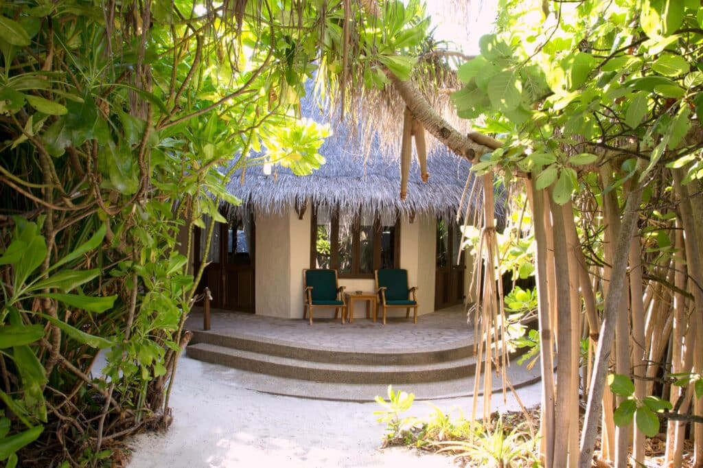 Entrée d’un bungalow entouré de végétation tropicale et de sable blanc