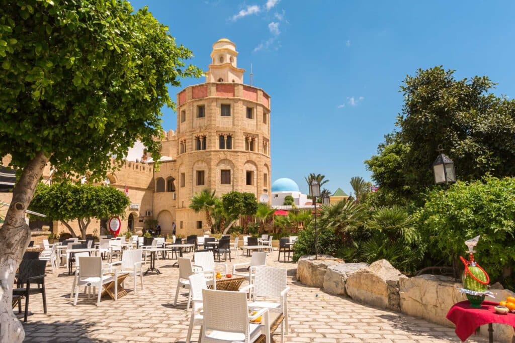 Terrasse ombragée d’un café en plein air face à un bâtiment en pierre de style tunisien