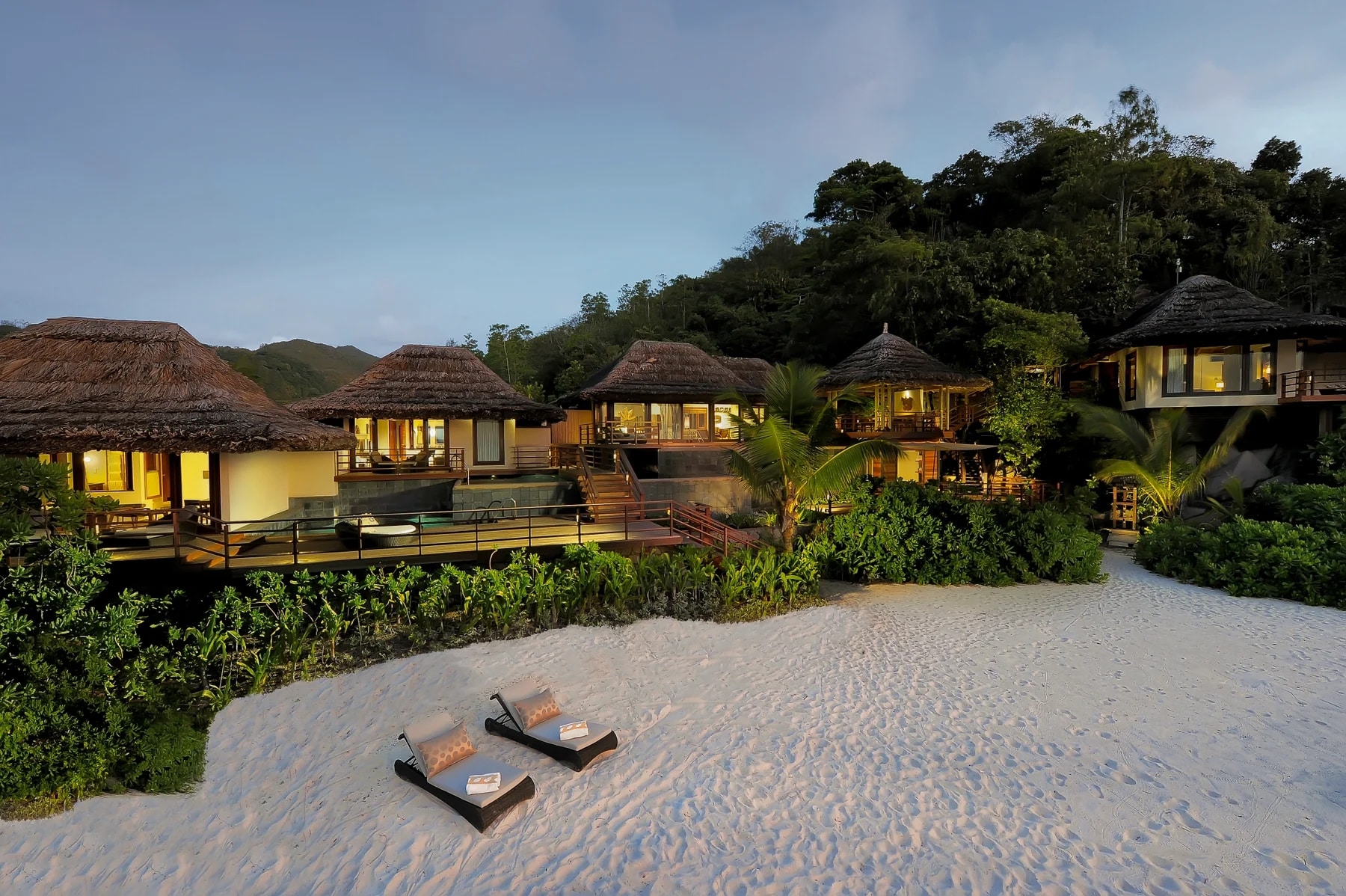 http://Villas%20de%20luxe%20avec%20toits%20de%20chaume%20en%20bord%20de%20plage%20au%20crépuscule,%20entourées%20de%20végétation%20tropicale.