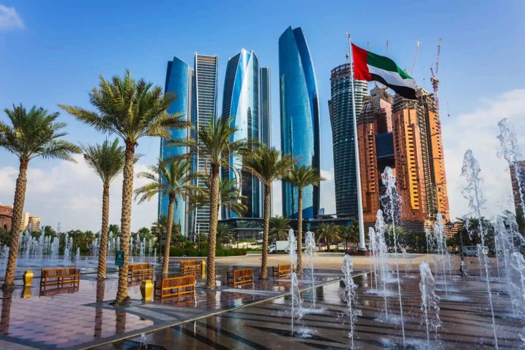 Vue des Etihad Towers à Abu Dhabi, avec fontaines, palmiers et drapeau des Émirats arabes unis au premier plan.