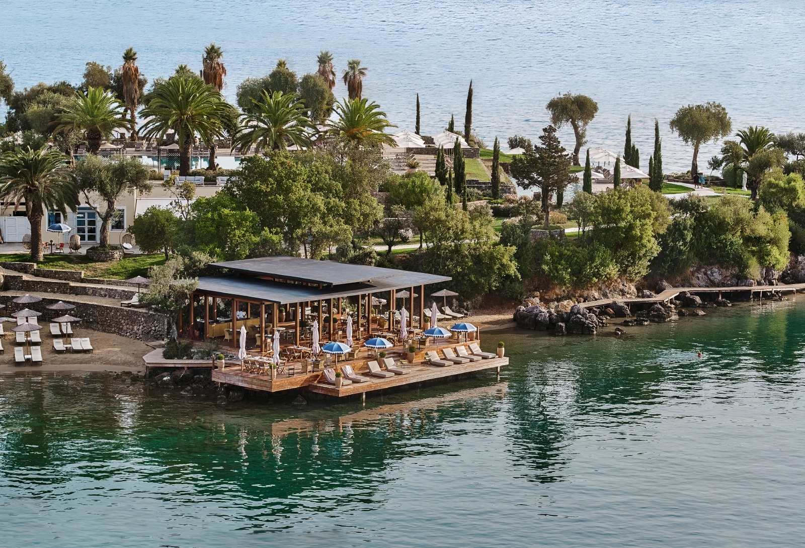 http://Restaurant%20en%20bois%20sur%20pilotis%20au%20Grecotel,%20en%20bord%20de%20mer,%20avec%20parasols%20bleus,%20transats%20et%20jardin%20méditerranéen.