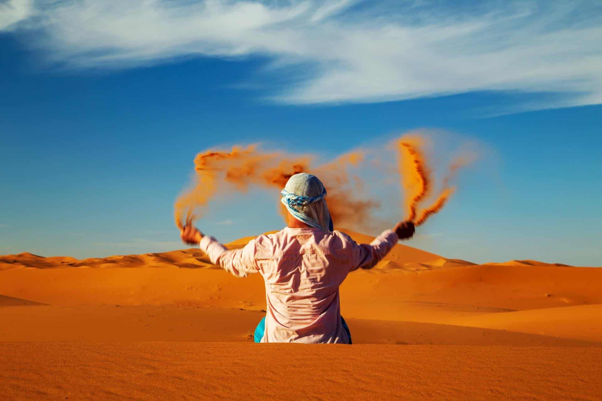 http://Homme%20vêtu%20d’un%20turban%20jouant%20avec%20le%20sable%20dans%20les%20dunes%20dorées%20du%20désert%20marocain