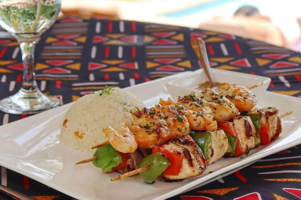 Brochettes de poulet et crevettes grillées servies avec du riz blanc parfumé, présentées dans une assiette sur une nappe africaine colorée.