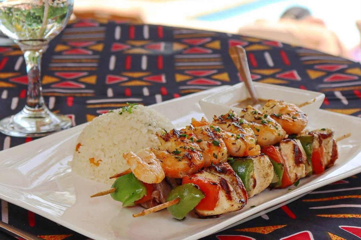 http://Brochettes%20de%20poulet%20et%20crevettes%20grillées%20servies%20avec%20du%20riz%20blanc%20parfumé,%20présentées%20dans%20une%20assiette%20sur%20une%20nappe%20africaine%20colorée.