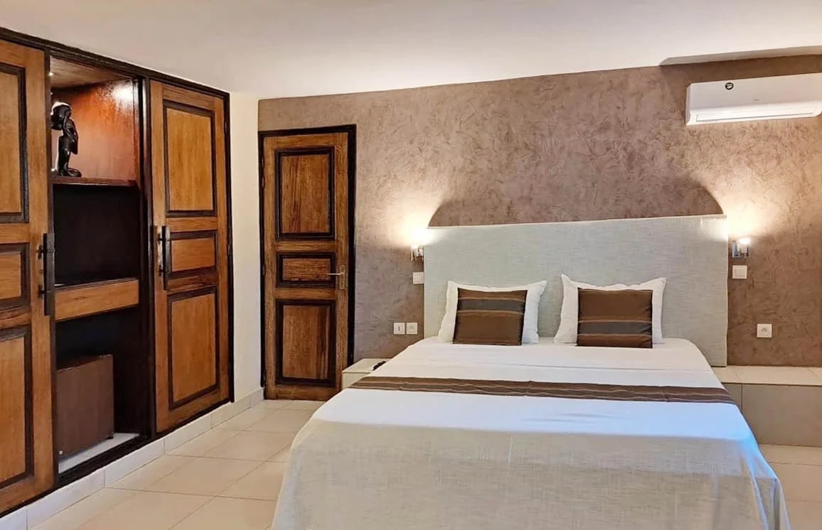 http://Chambre%20d’hôtel%20confortable%20avec%20lit%20double,%20climatisation%20murale%20et%20décoration%20en%20bois%20au%20style%20africain.