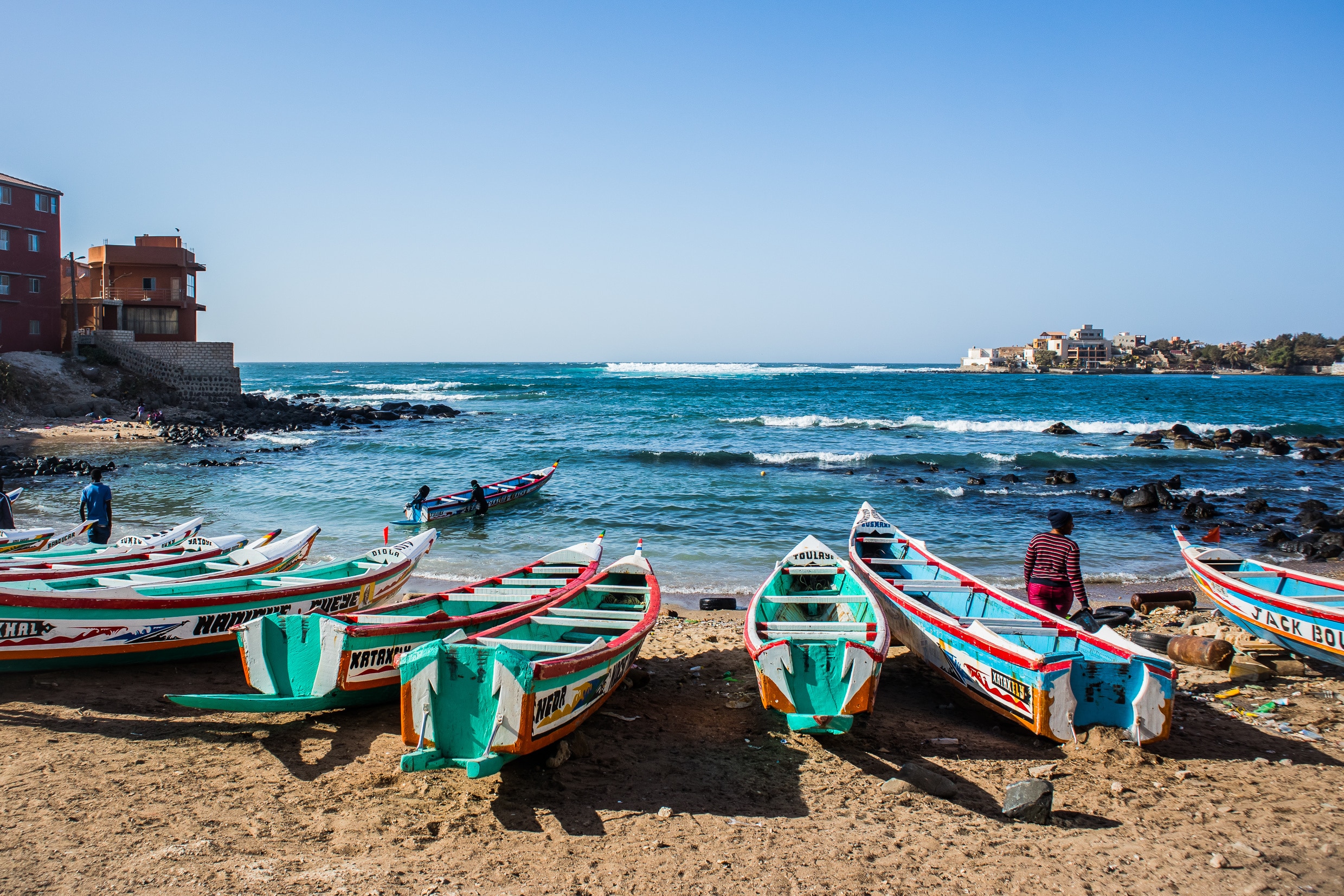 http://Pirogues%20colorées%20de%20pêcheurs%20alignées%20sur%20une%20plage%20rocheuse%20à%20Dakar,%20avec%20vue%20sur%20l’île%20de%20Gorée.