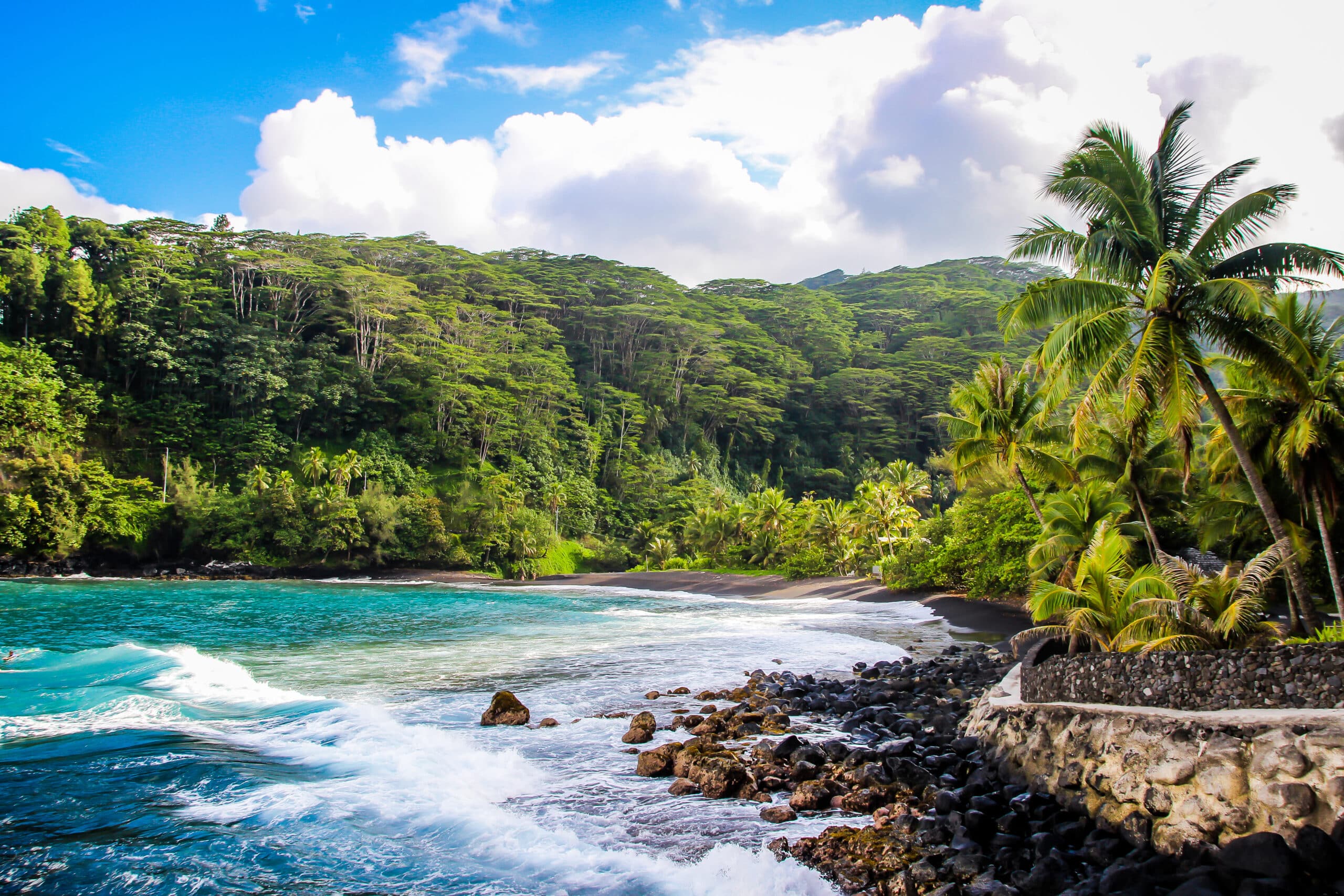 http://Vagues%20s’écrasant%20sur%20des%20rochers%20volcaniques%20face%20à%20une%20plage%20de%20sable%20noir,%20entourée%20d’une%20végétation%20dense.