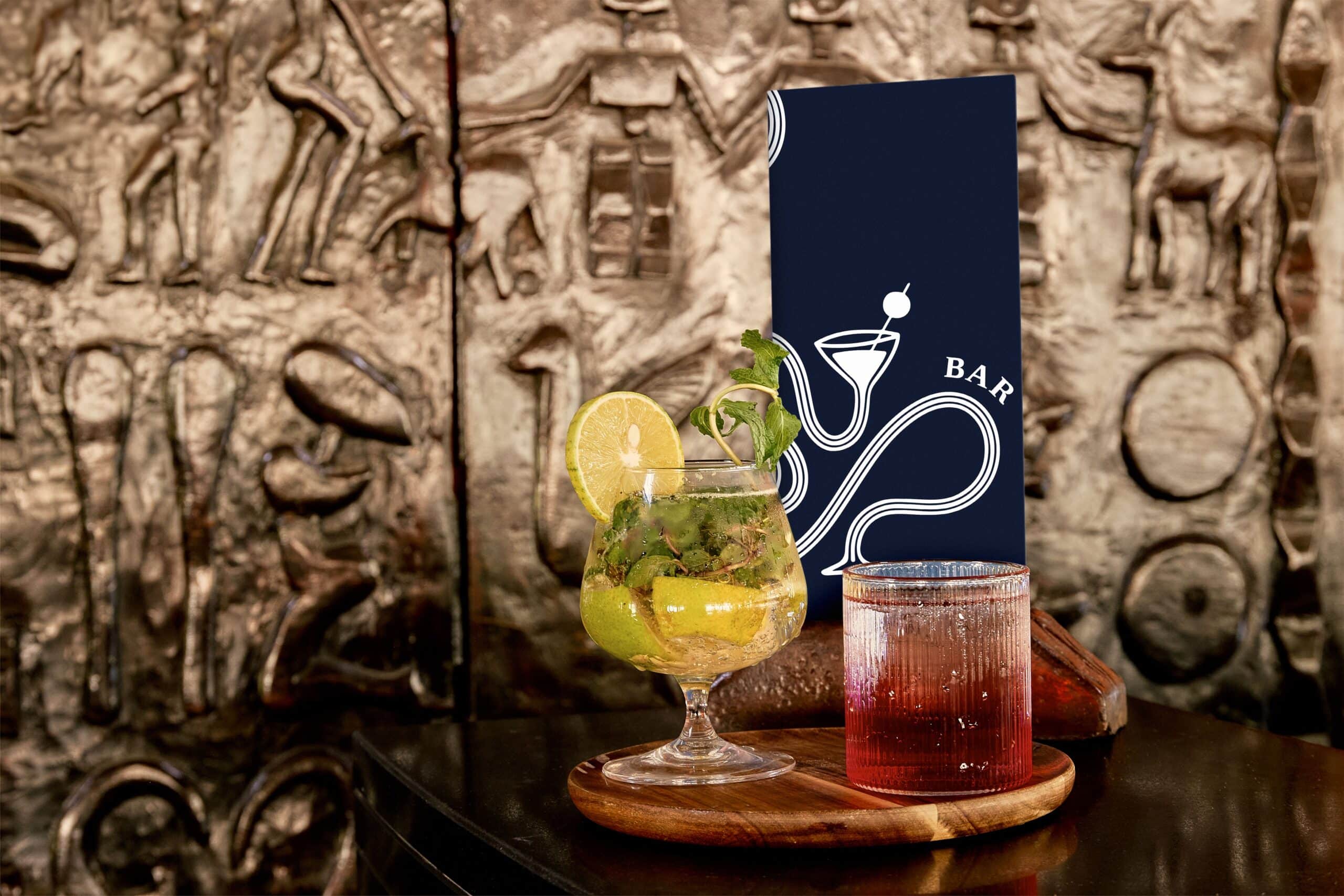 http://Deux%20cocktails%20colorés%20sur%20un%20comptoir%20avec%20menu%20du%20bar%20en%20fond,%20décor%20mural%20africain