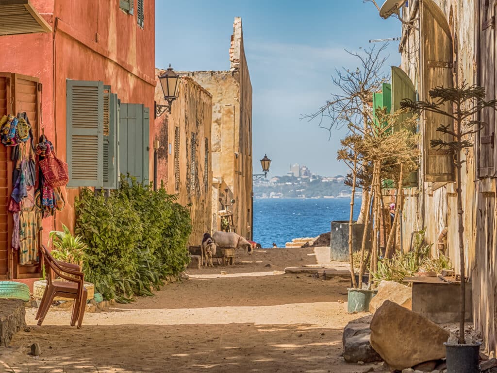 Ruelle sableuse bordée de maisons coloniales colorées sur l’île de Gorée, avec vue sur l’océan Atlantique.