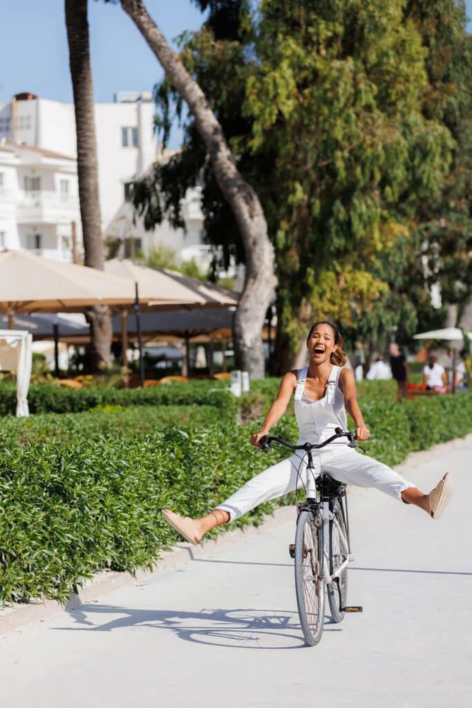 Femme souriante en blanc faisant du vélo sur une promenade bordée de verdure dans une station balnéaire.