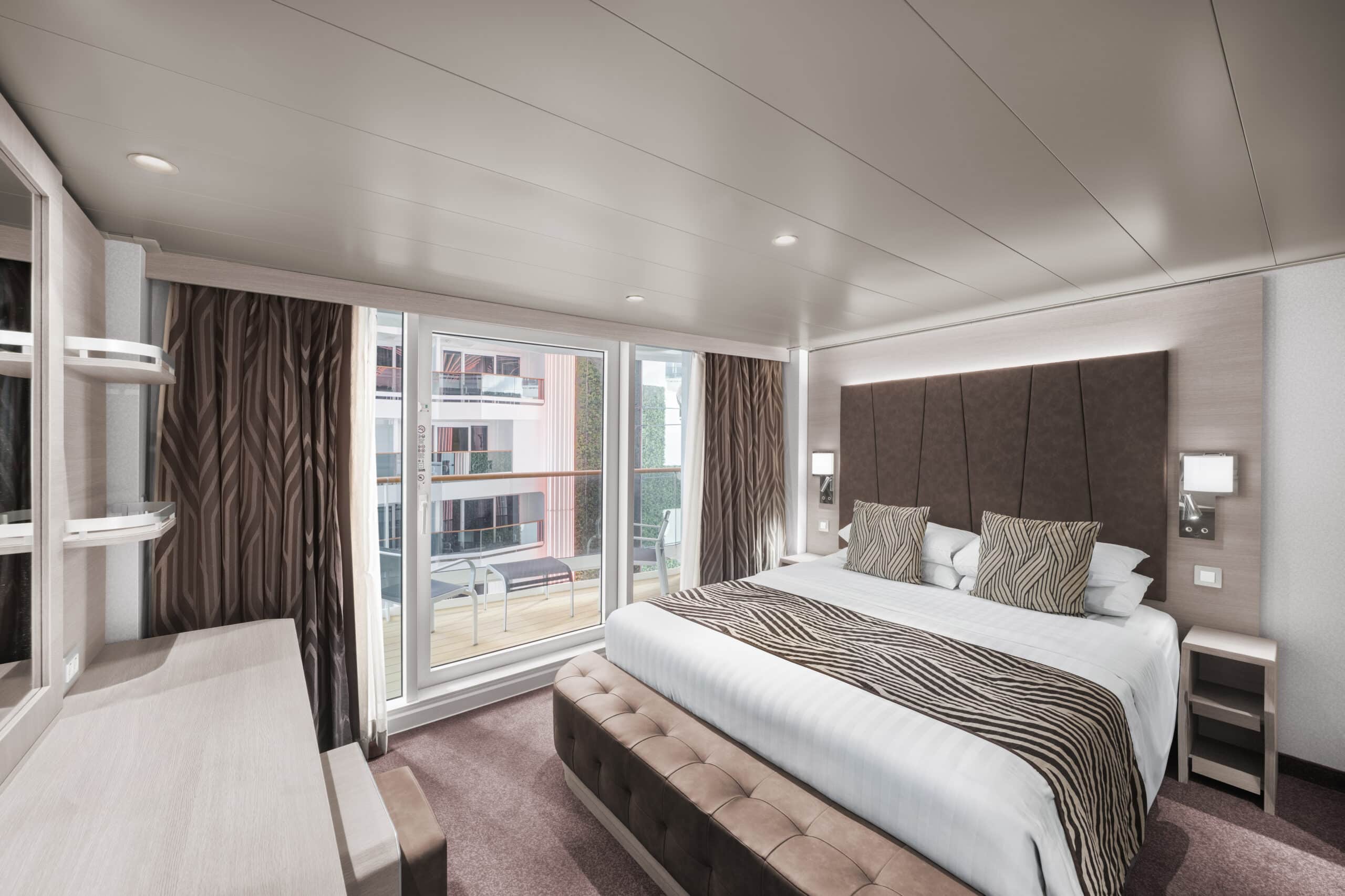 Cabine avec balcon d'un navire de croisière, équipée d'un lit double, de coussins à motifs et d'une baie vitrée donnant sur un balcon aménagé.