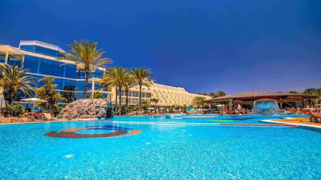 Grande piscine extérieure entourée de palmiers avec bar central dans un hôtel en bord de mer.