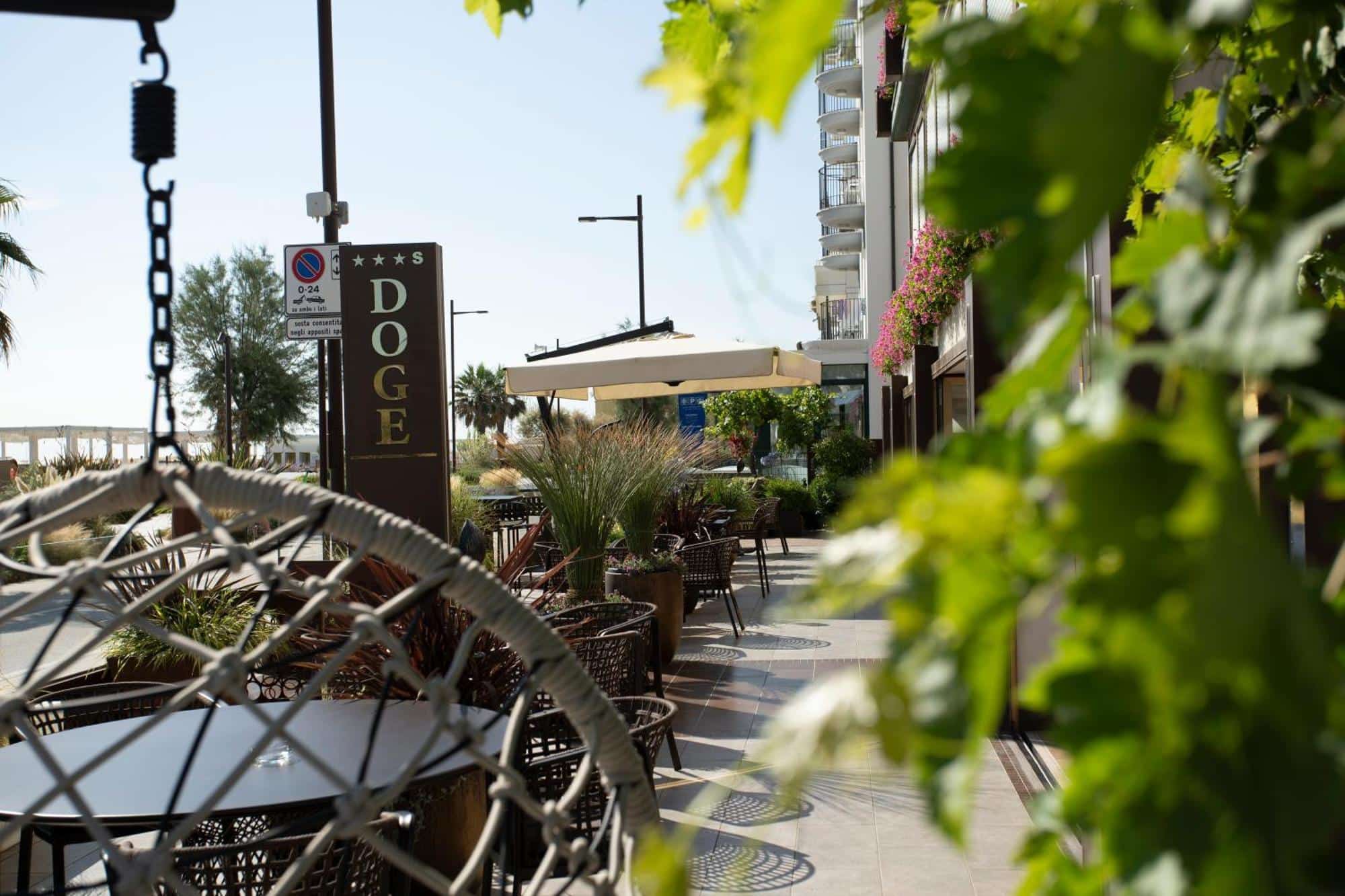 http://Terrasse%20fleurie%20de%20l’hôtel%20Doge%20avec%20tables,%20chaises%20et%20parasols%20sous%20un%20ciel%20ensoleillé.