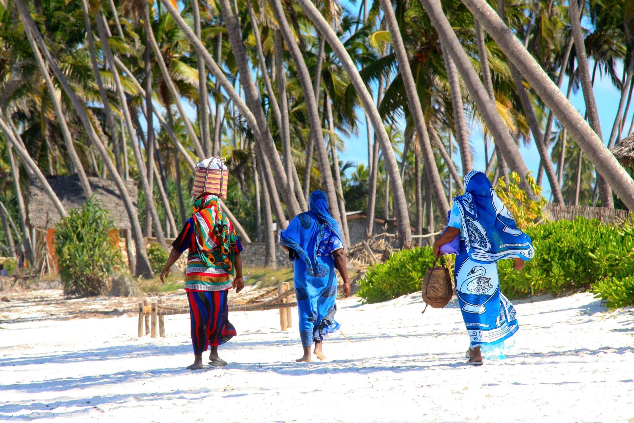 http://Trois%20femmes%20en%20kanga%20coloré%20marchant%20sur%20une%20plage%20bordée%20de%20palmiers%20à%20Zanzibar