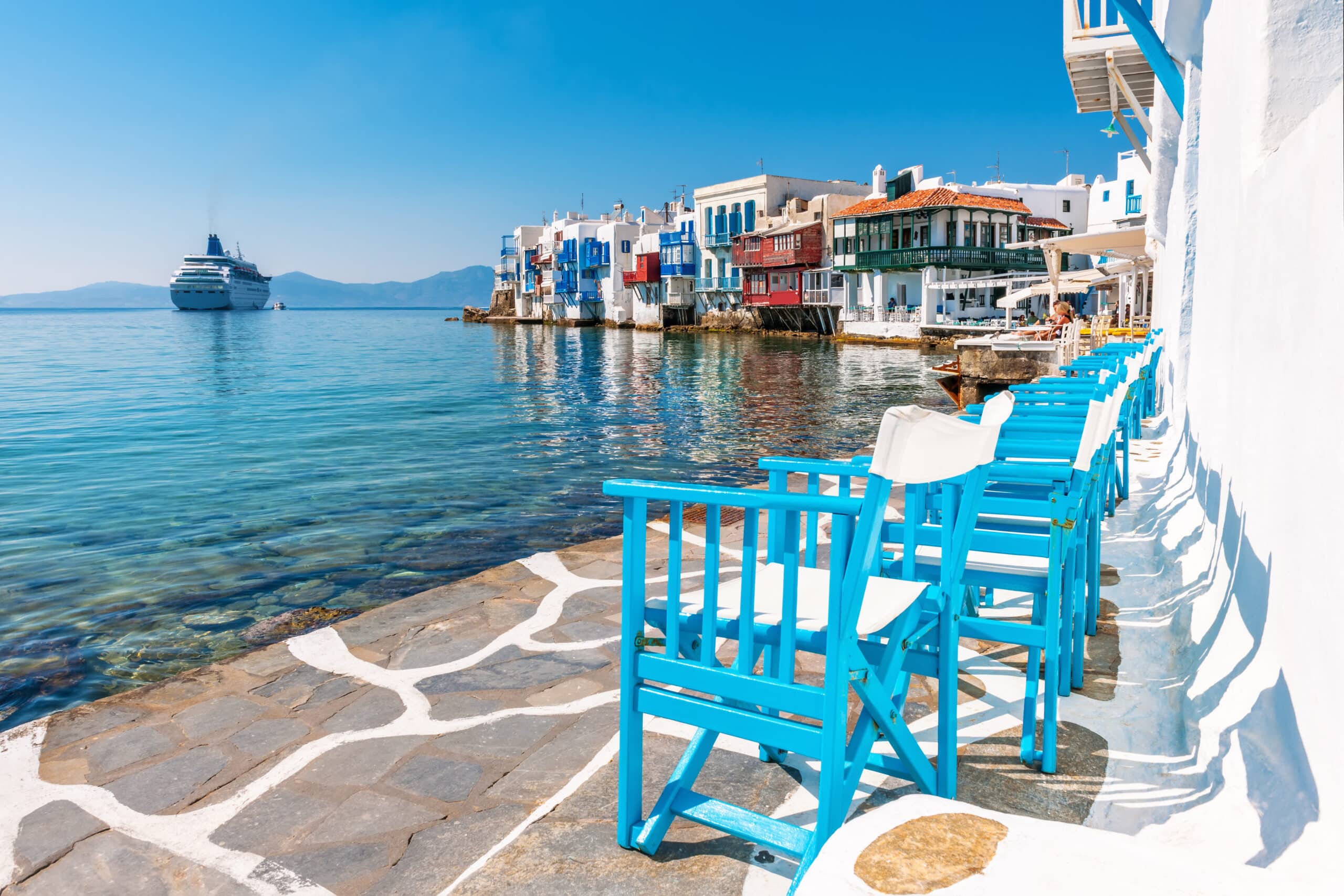 http://Terrasse%20en%20bord%20de%20mer%20avec%20chaises%20bleues%20à%20Mykonos%20et%20vue%20sur%20les%20maisons%20colorées%20de%20Petite%20Venise.