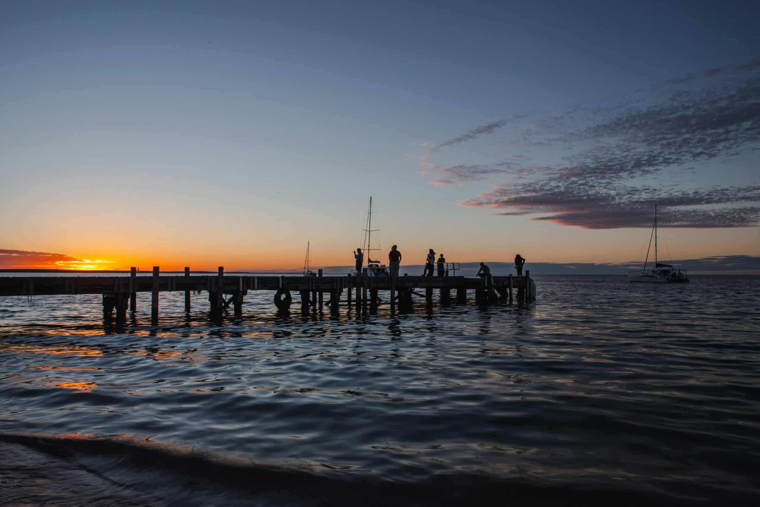 http://Silhouettes%20de%20voyageurs%20sur%20un%20ponton%20au%20coucher%20du%20soleil%20avec%20voiliers%20sur%20la%20mer