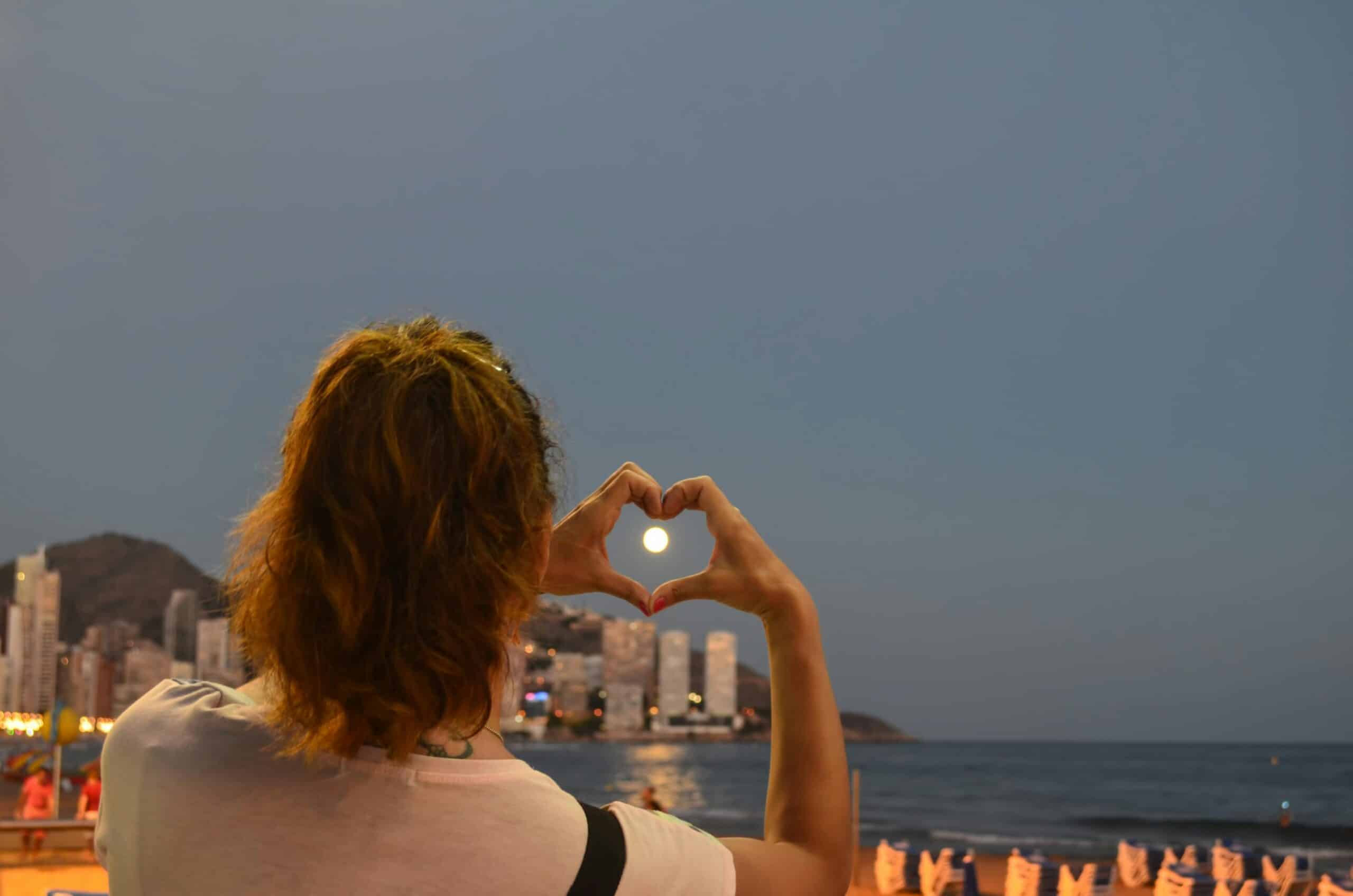 http://Personne%20formant%20un%20cœur%20avec%20ses%20mains%20encadrant%20la%20lune%20au-dessus%20de%20la%20mer%20et%20de%20la%20ville