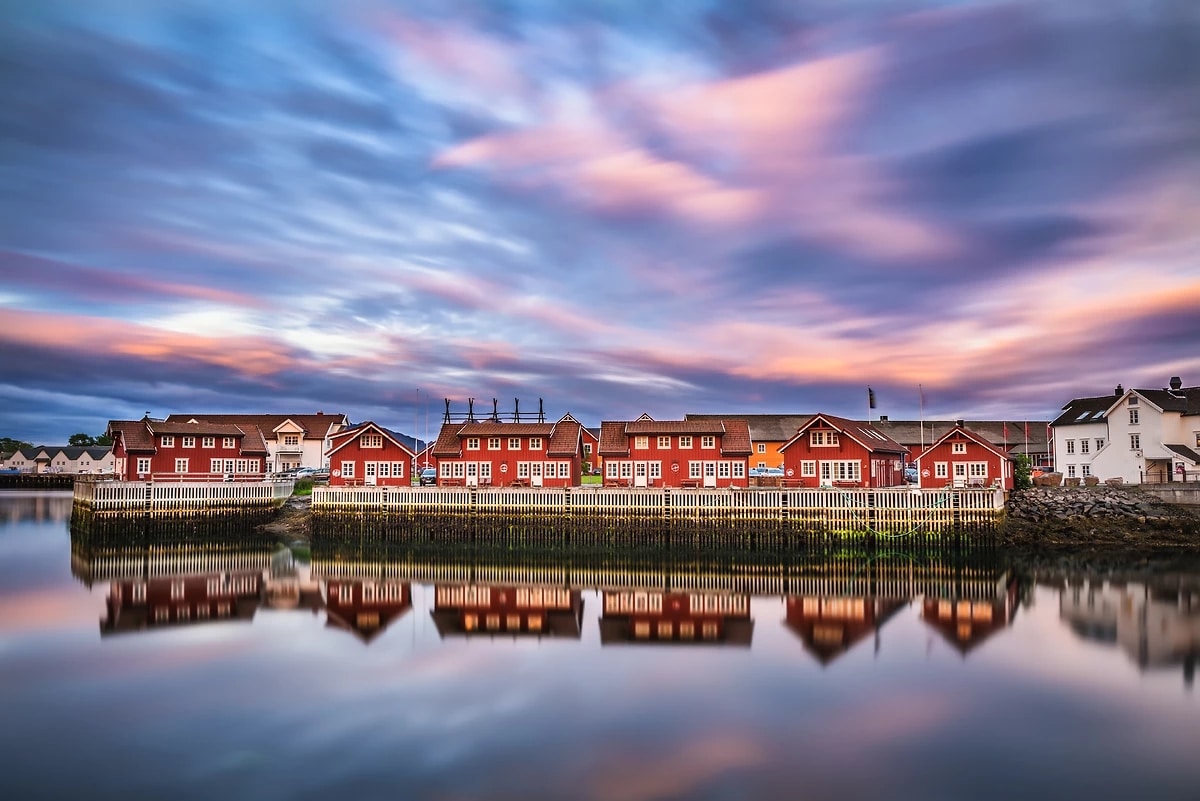 http://Maisons%20rouges%20norvégiennes%20au%20bord%20de%20l’eau%20avec%20reflets%20et%20ciel%20coloré