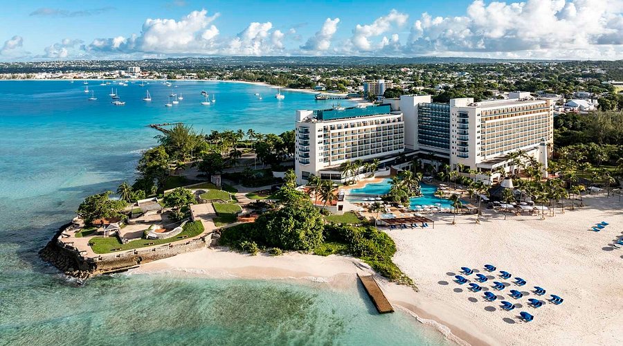 Vue aérienne d’un resort en bord de mer avec piscines, plage de sable blanc et eaux turquoise.