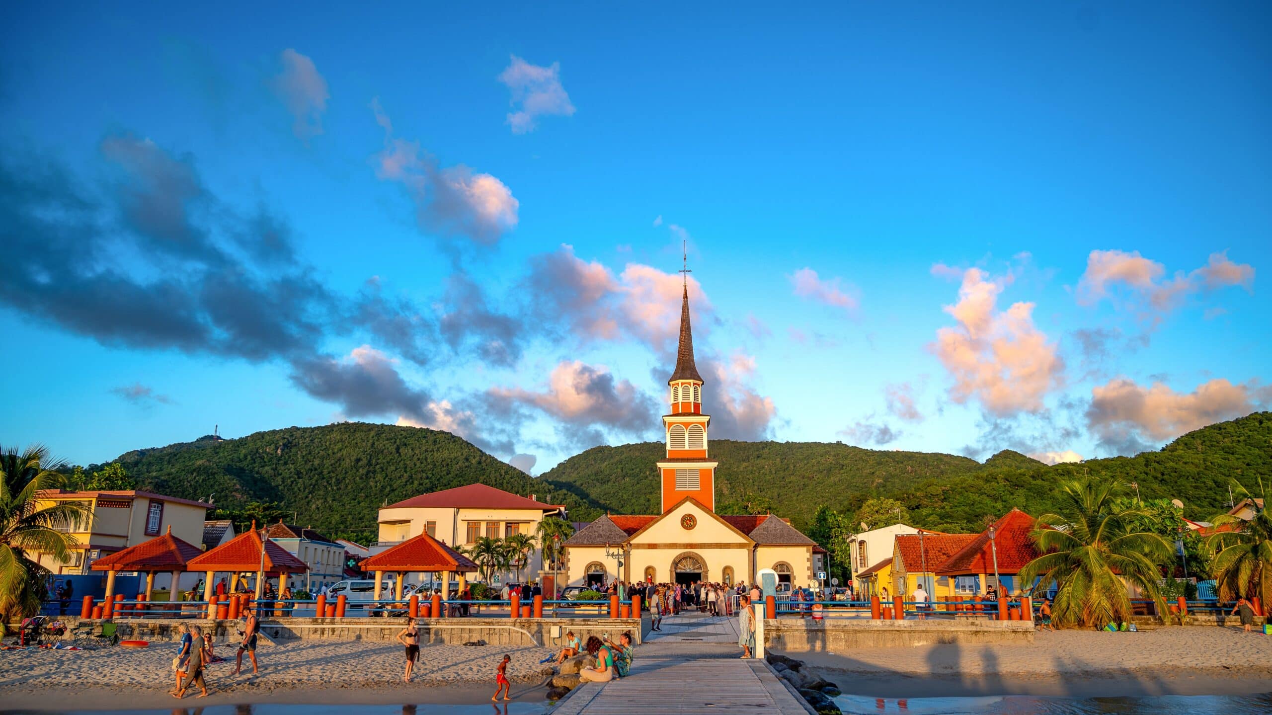 http://Église%20au%20cœur%20d'un%20village%20côtier%20martiniquais%20avec%20plage,%20promenade%20et%20montagnes%20verdoyantes%20en%20arrière-plan.