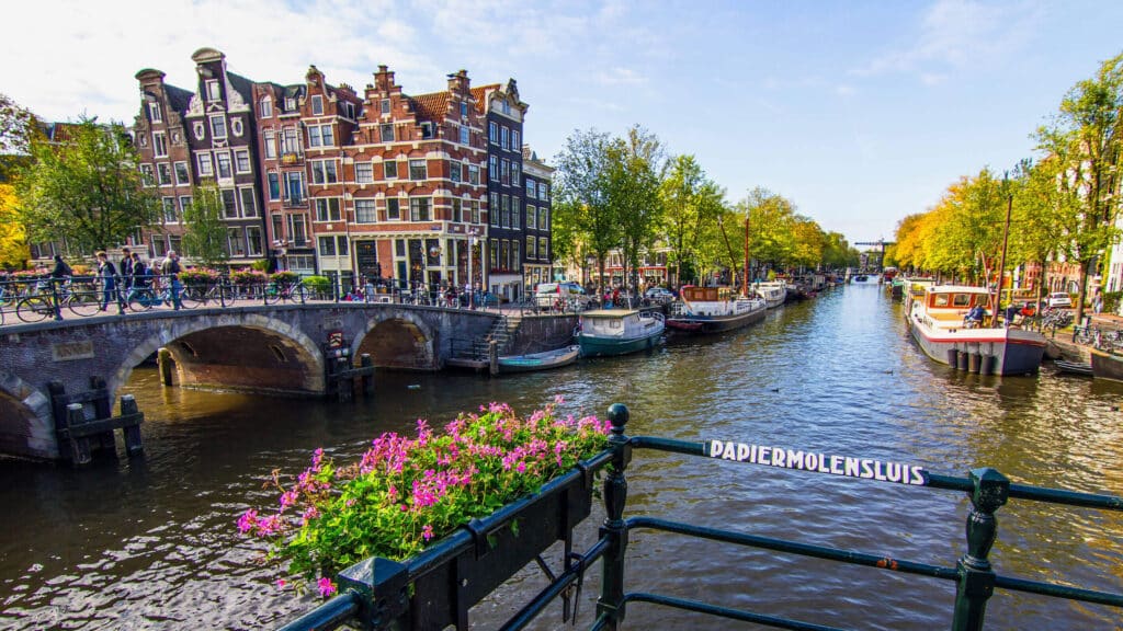 Canal d’Amsterdam bordé de maisons traditionnelles, de bateaux et d’arbres avec un pont en pierre.