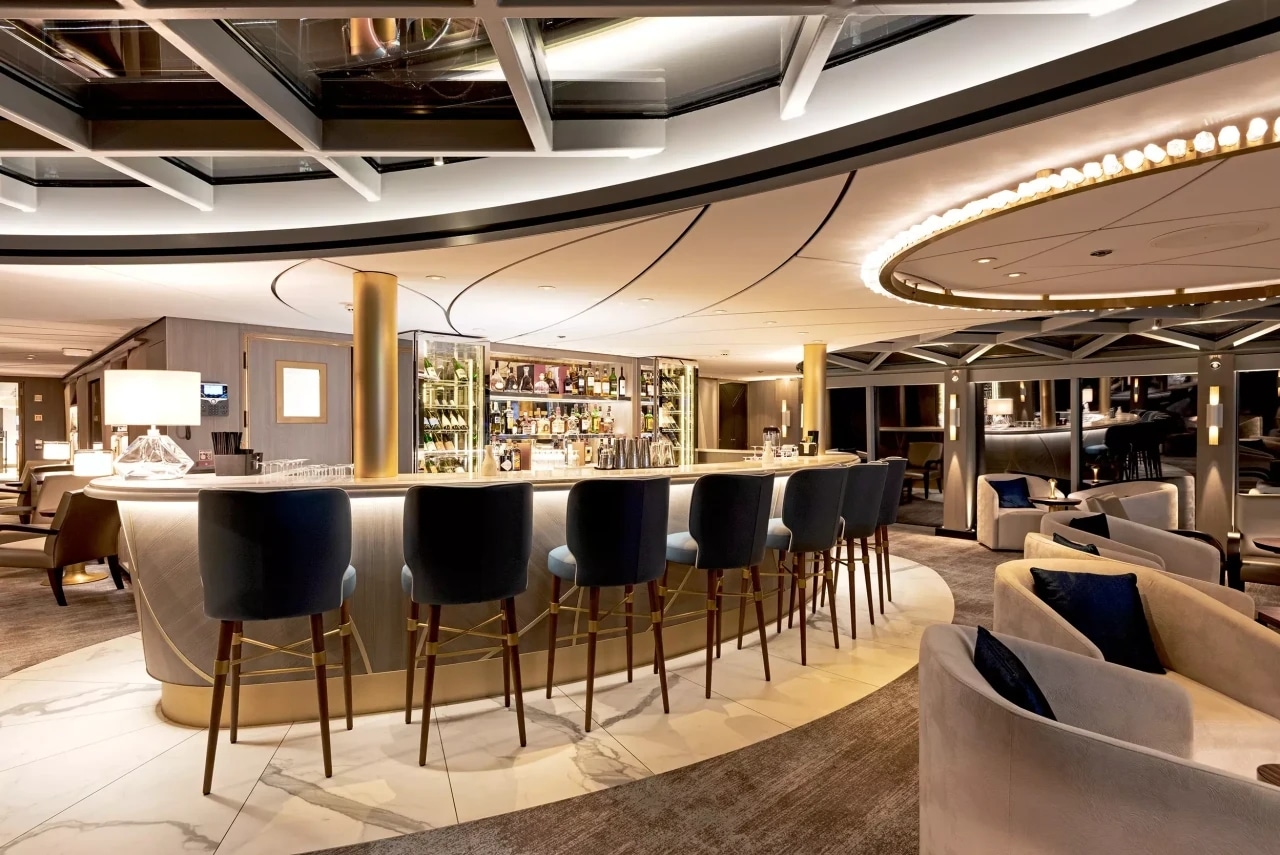 Bar lounge élégant à bord d’un bateau de croisière fluviale avec comptoir central, fauteuils et tabourets.