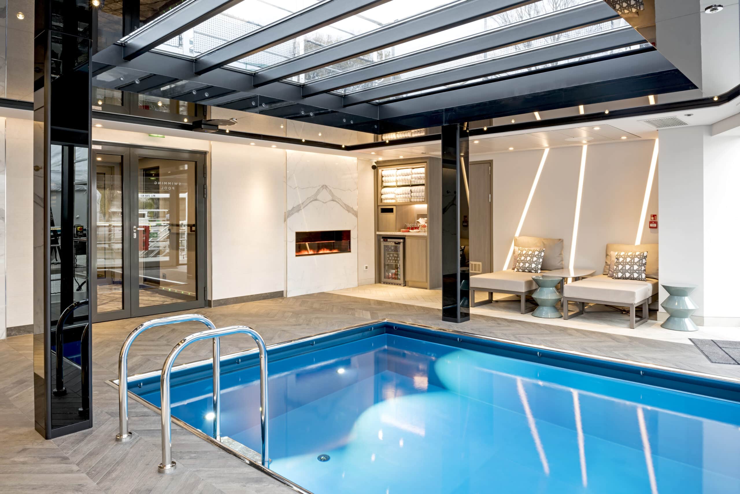 Piscine intérieure avec verrière, cheminée et espace de détente équipé de transats.