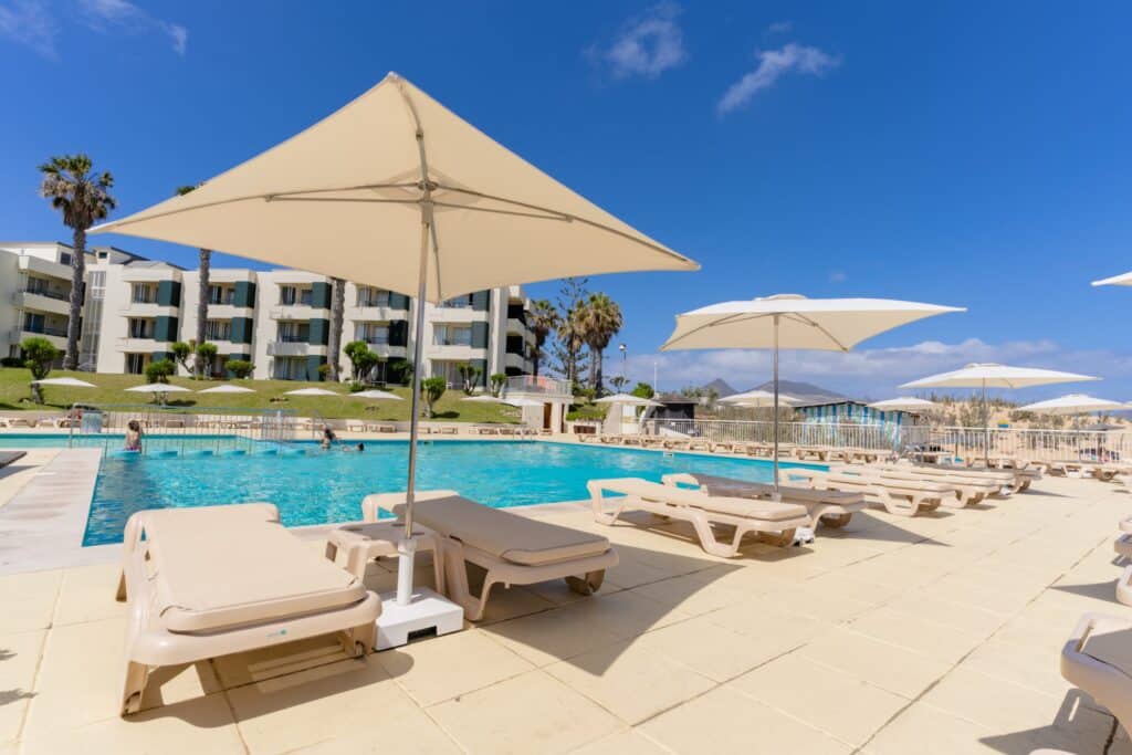 Piscine extérieure d’un hôtel à Porto Santo entourée de transats, parasols et palmiers sous un ciel bleu.