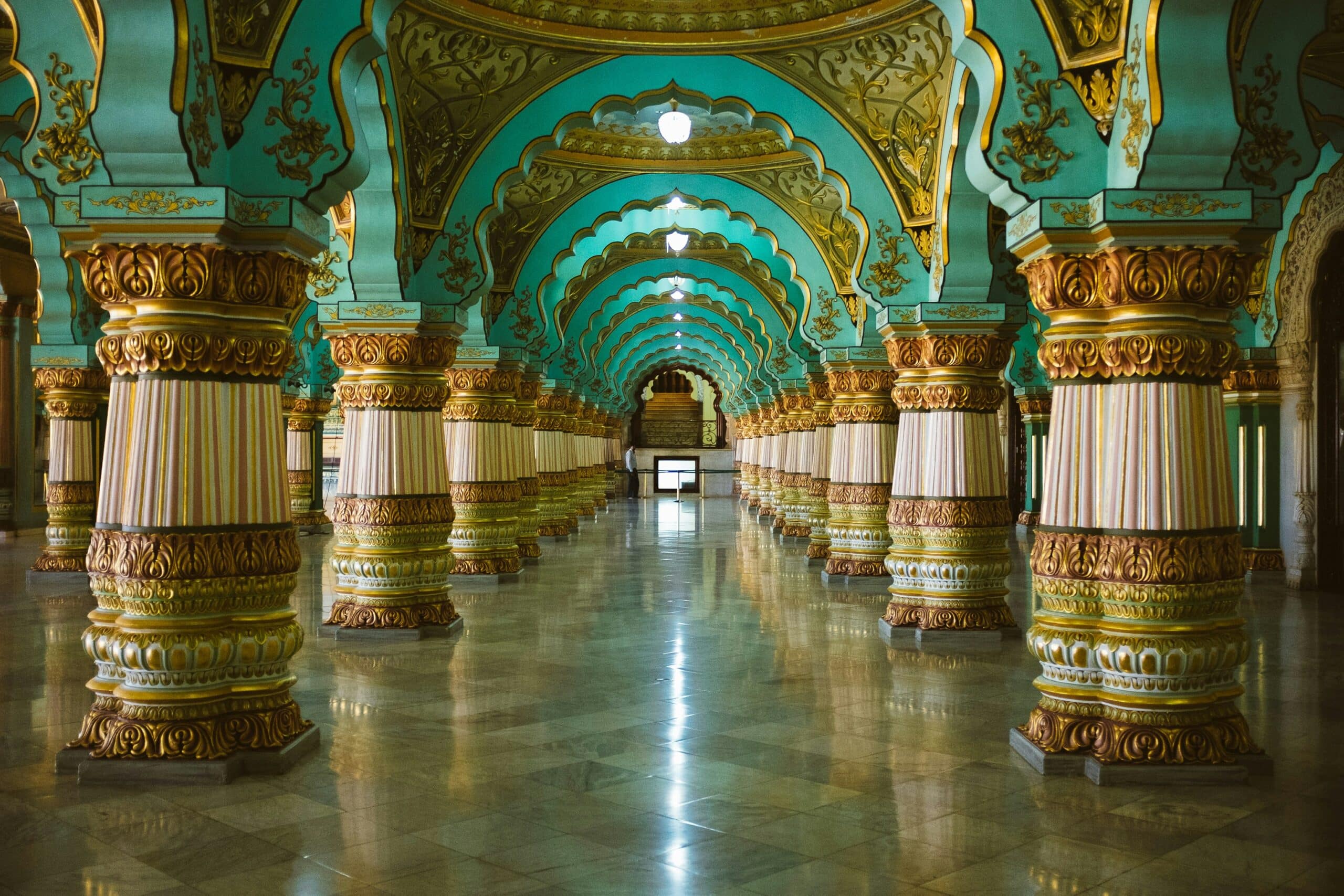 http://Galerie%20intérieure%20du%20palais%20de%20Mysore%20ornée%20de%20colonnes%20dorées%20et%20d'arches%20turquoise%20richement%20décorées.