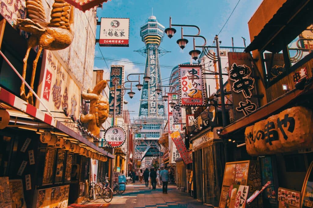 Rue animée du quartier Shinsekai à Osaka avec ses enseignes lumineuses, ses restaurants et la tour Tsutenkaku en arrière-plan.