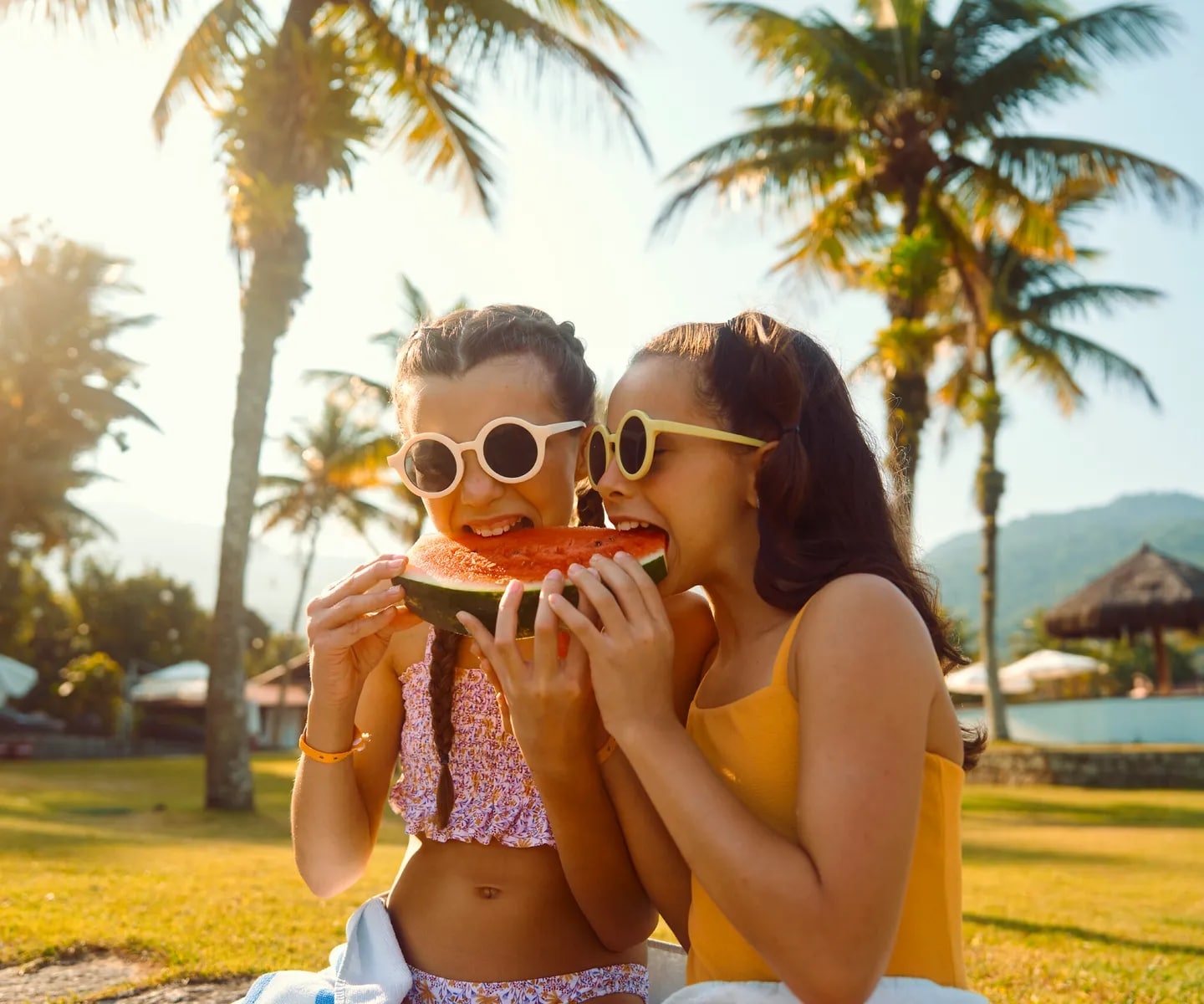 http://Deux%20jeunes%20filles%20partageant%20une%20tranche%20de%20pastèque%20dans%20le%20jardin%20d’un%20hôtel%20tropical%20bordé%20de%20palmiers.