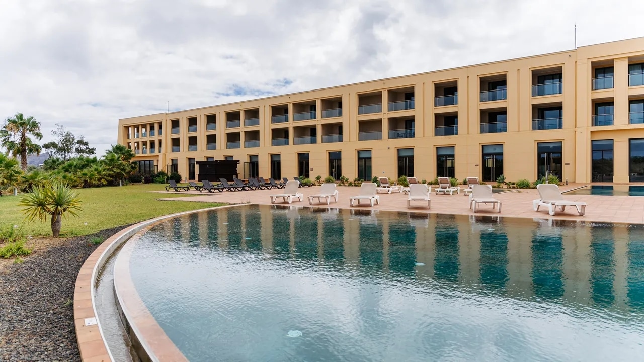 http://Grande%20piscine%20extérieure%20d’un%20hôtel%20à%20Porto%20Santo%20avec%20transats,%20jardin%20et%20chambres%20avec%20balcon.